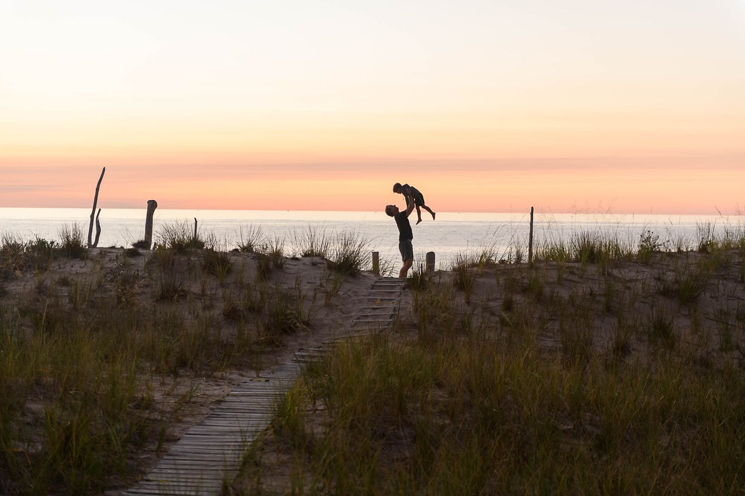 silhouette of boy and man in front of golden sky and water on a path with dune grass on either side