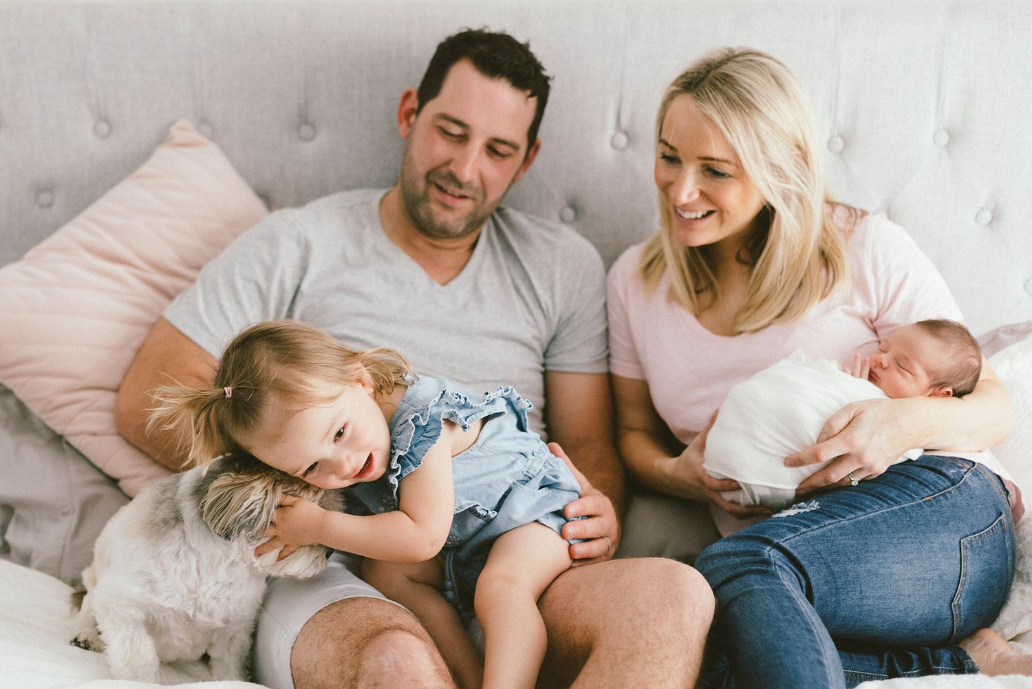 Family relaxing together on gray upholstered bed with pink pillows.