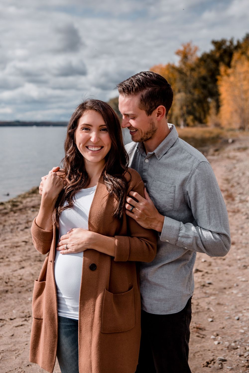 Romantic couple photos on a beach with autumn trees and cloudy skies.