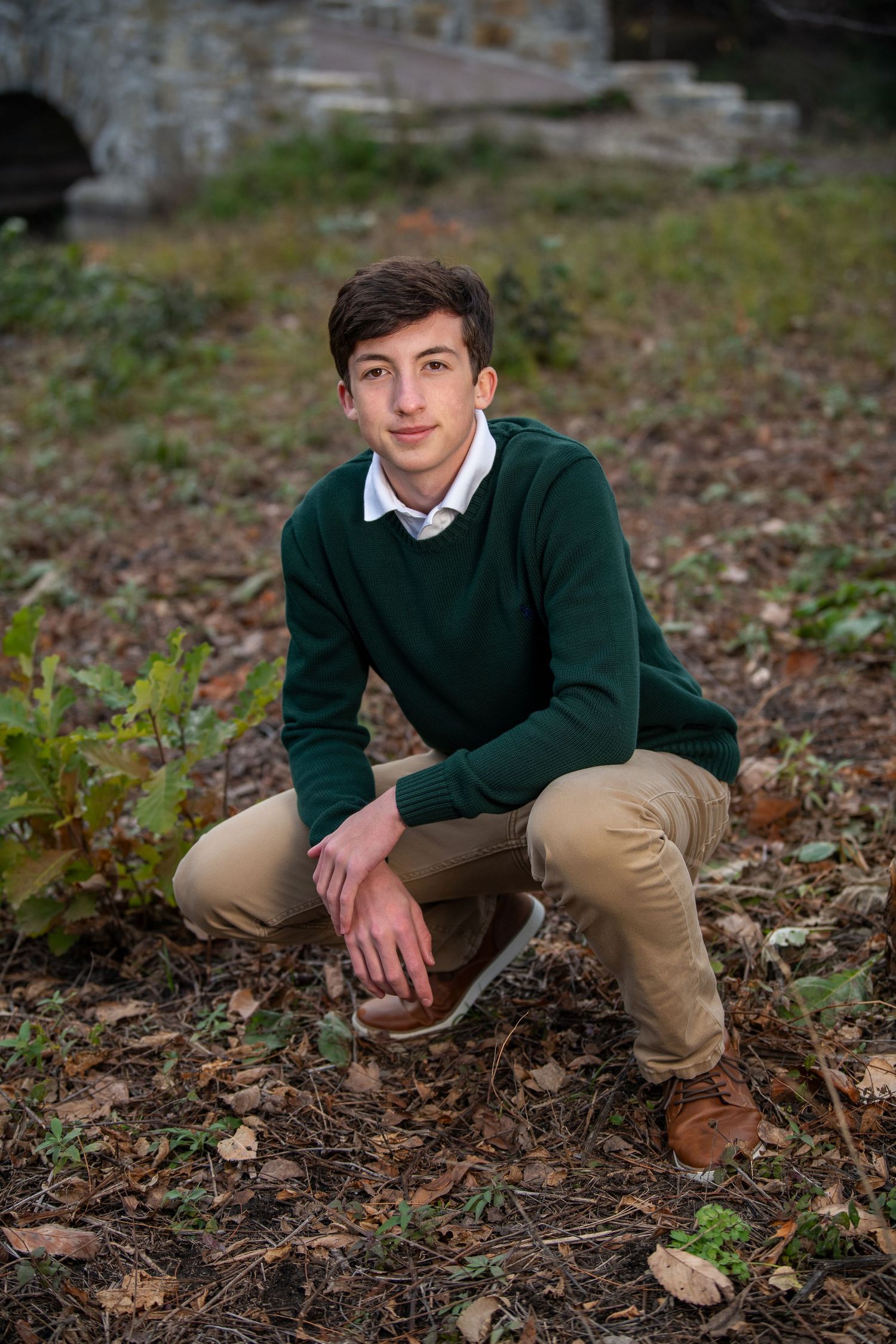 Student in green sweater and khakis crouches in autumn leaves for outdoor portrait.