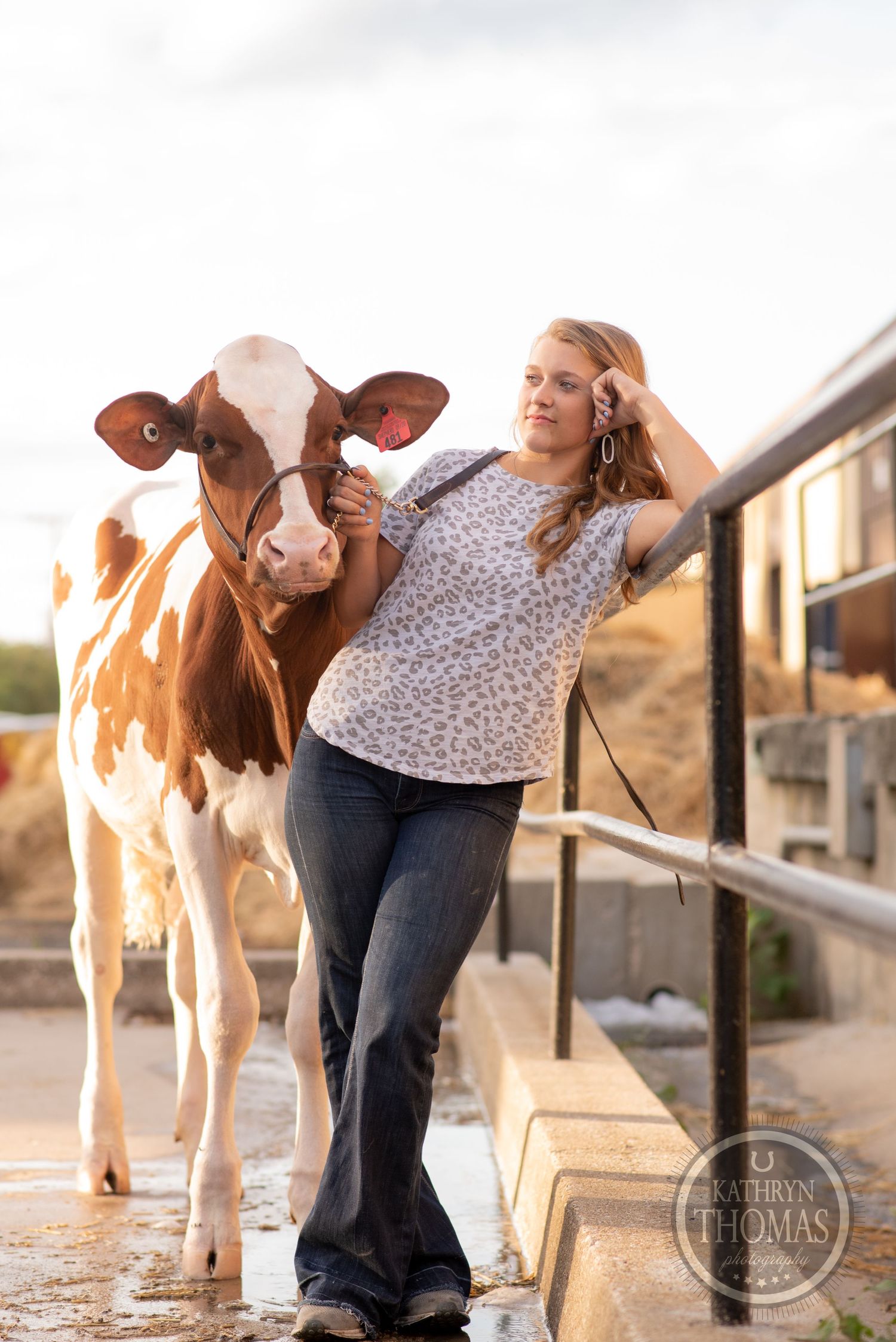 A brown and white dairy cow poses with a person in a patterned shirt and jeans on a sunny farm day.