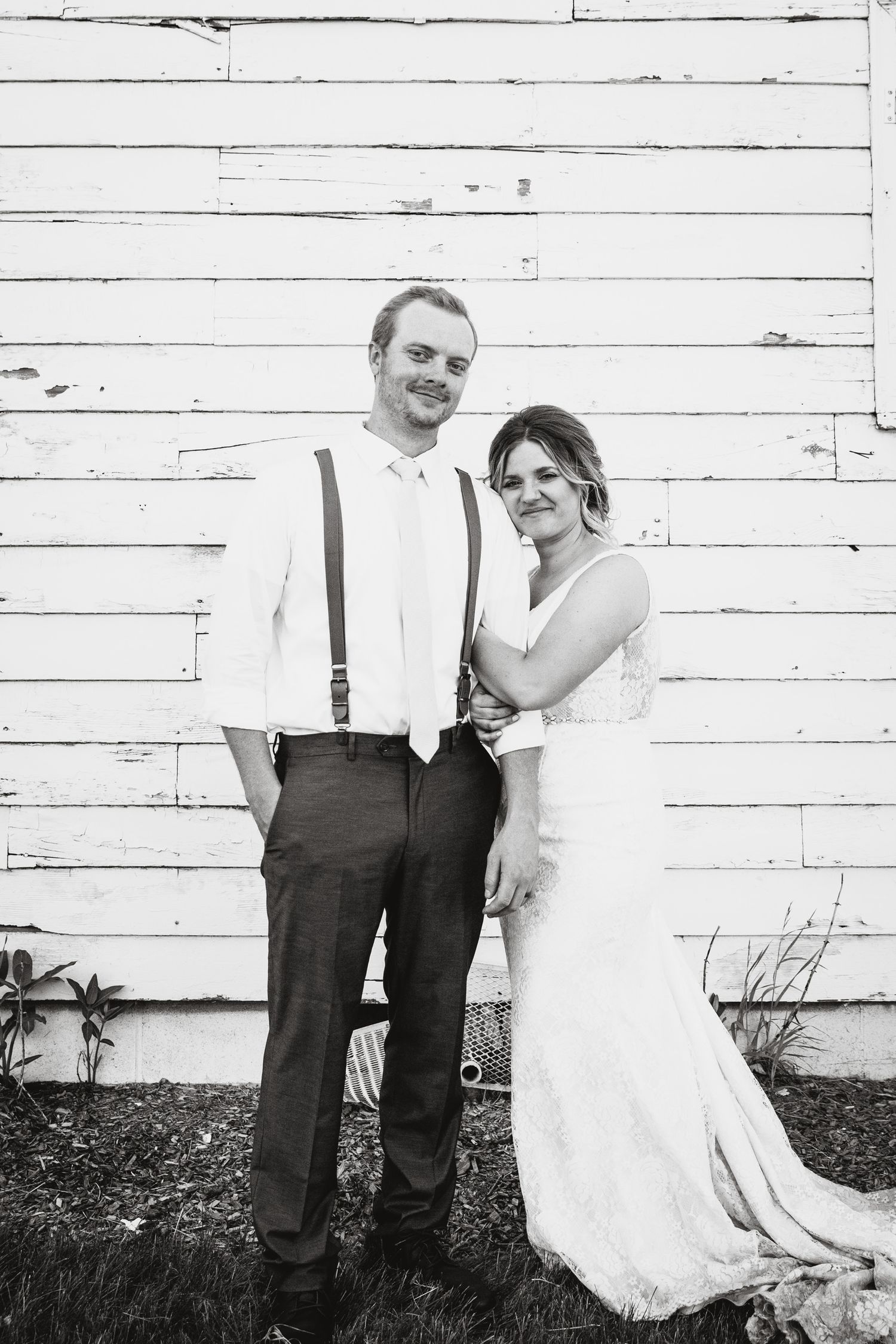 Black-and-white portrait of a couple on their wedding day. A rustic white barn frames the couple as a backdrop.