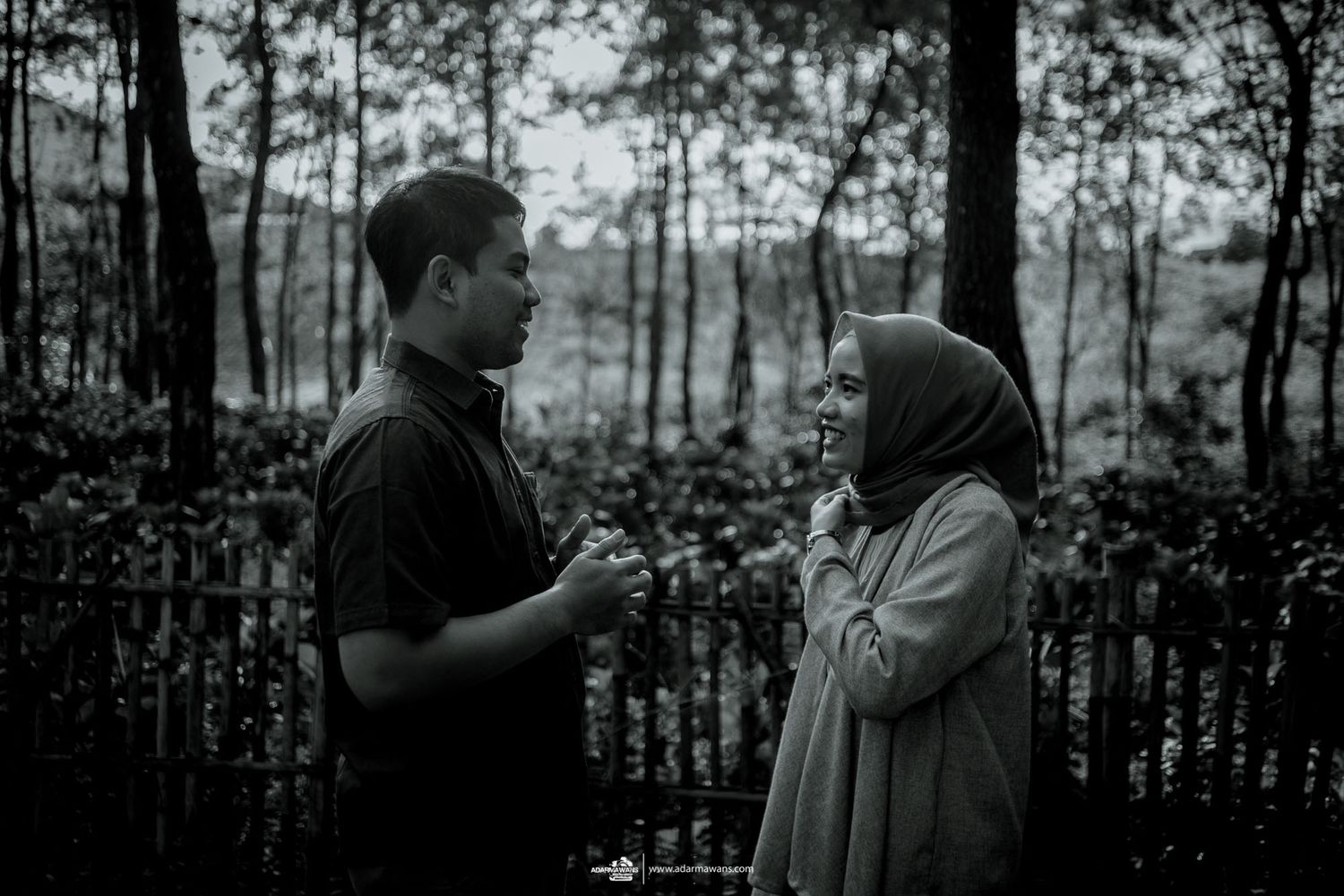 A couple shares an intimate conversation in a moody forest setting with trees in the background.