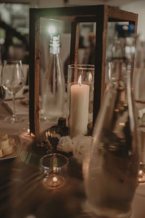 Elegant table setting with pillar candle in glass lantern surrounded by wine glasses and bottles.