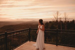 Bride in flowing white dress admiring mountain sunset view from wooden deck.
