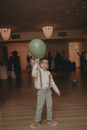 Young boy in suspenders holds a green balloon on a dance floor.