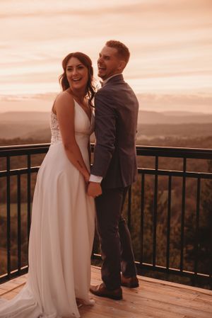 Joyful wedding couple sharing laughter on mountain overlook during sunset.