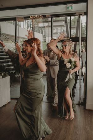 Wedding party in olive green dresses dancing joyfully during reception celebration.