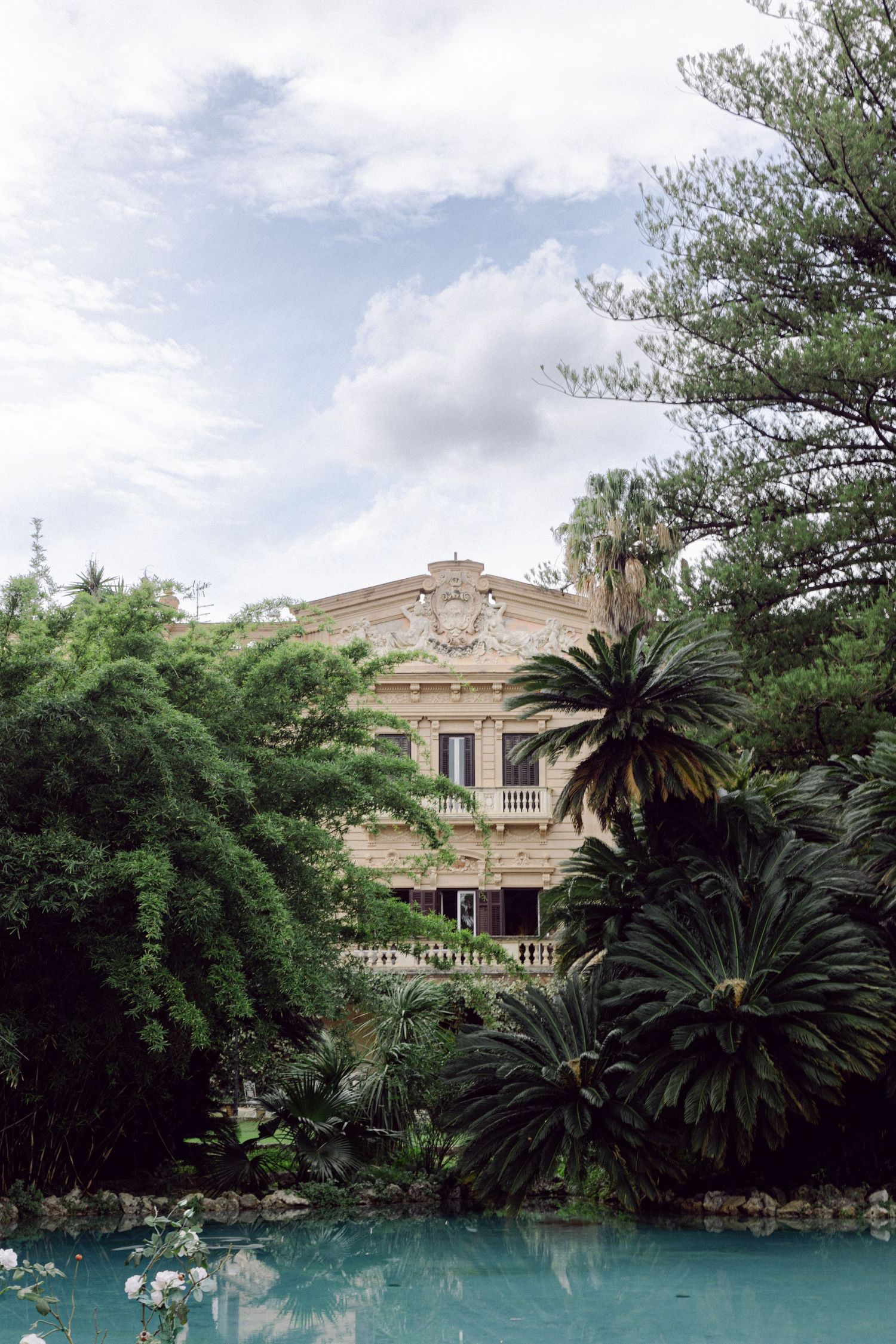 Elegant mansion with palm trees and a reflecting pool under a cloudy sky.