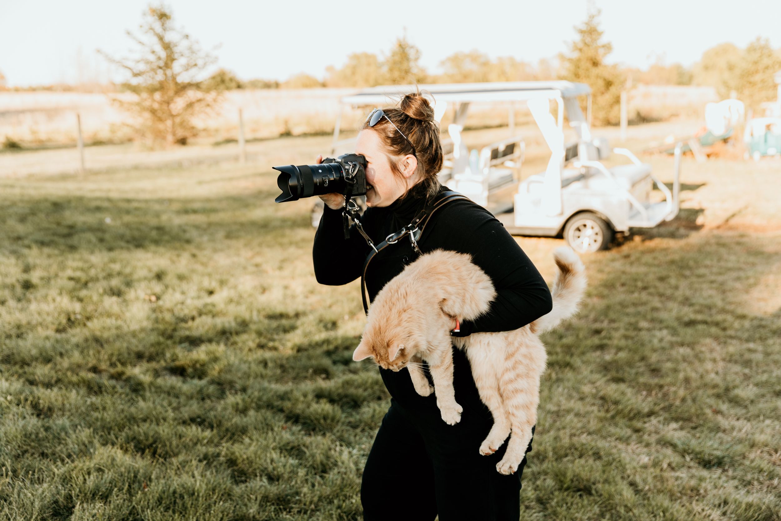 Photographer wearing black holds camera while carrying golden retriever puppy outdoors.