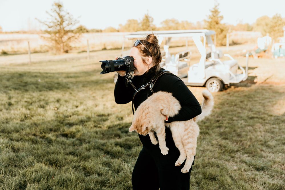 Photographer wearing black holds camera while carrying golden retriever puppy outdoors.