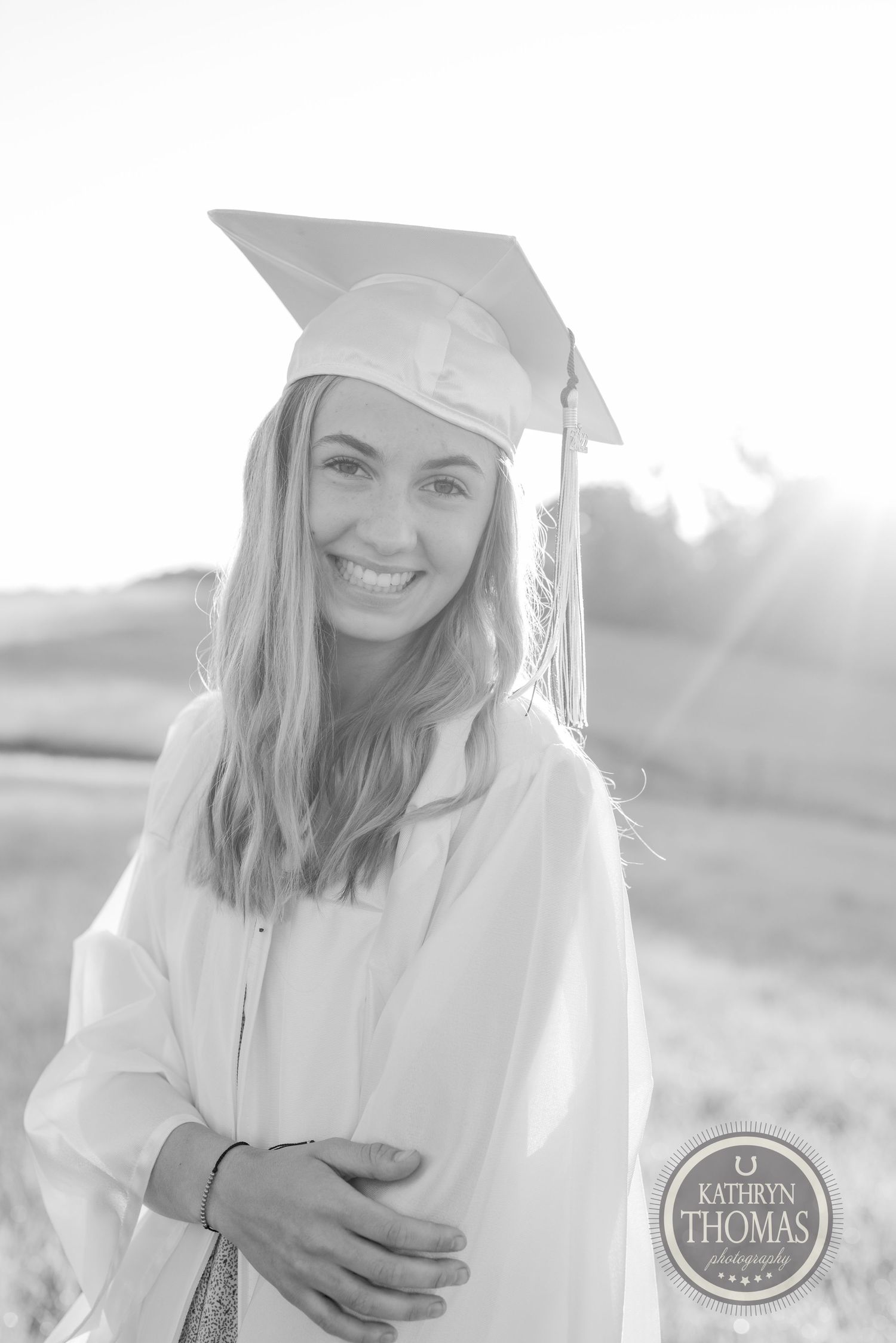 Black and white graduation portrait showing graduate in cap and gown against rural landscape.