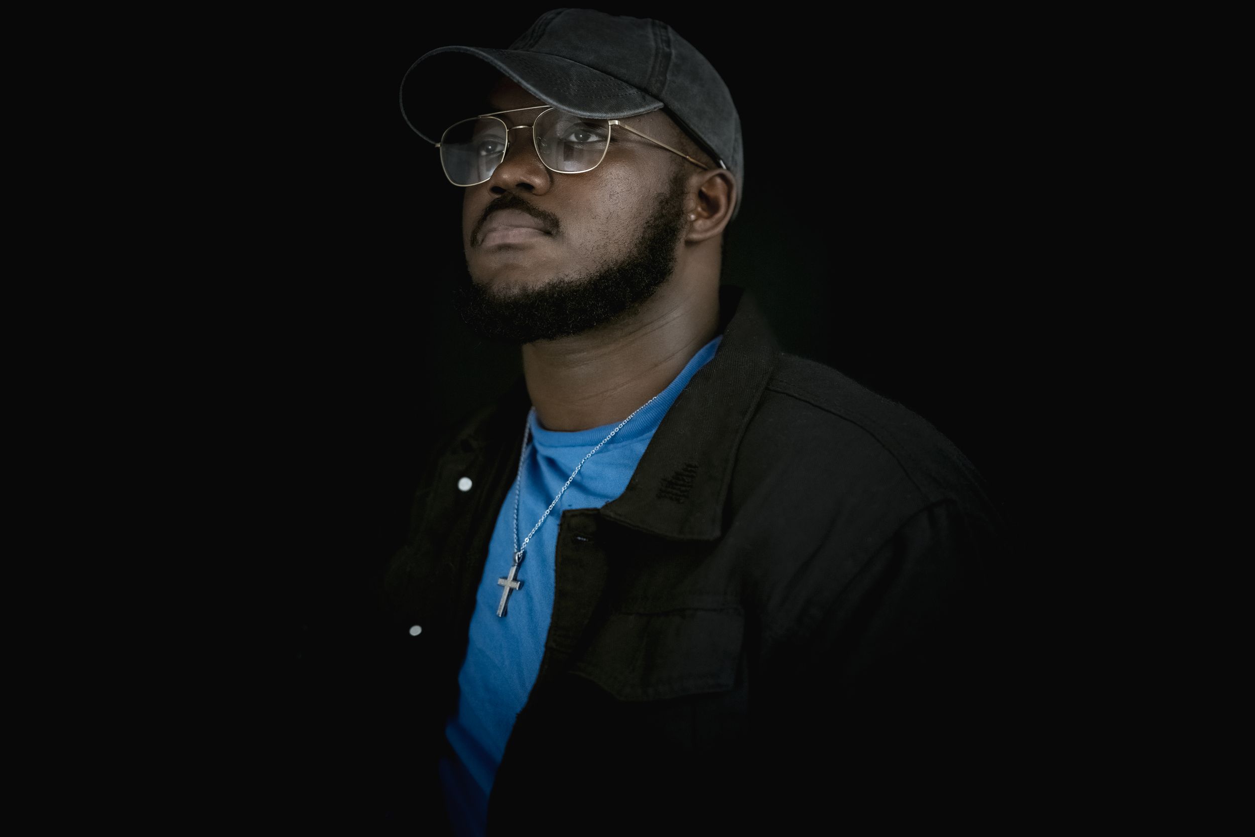 A person in glasses and cap looks upward while wearing a blue shirt and dark jacket.