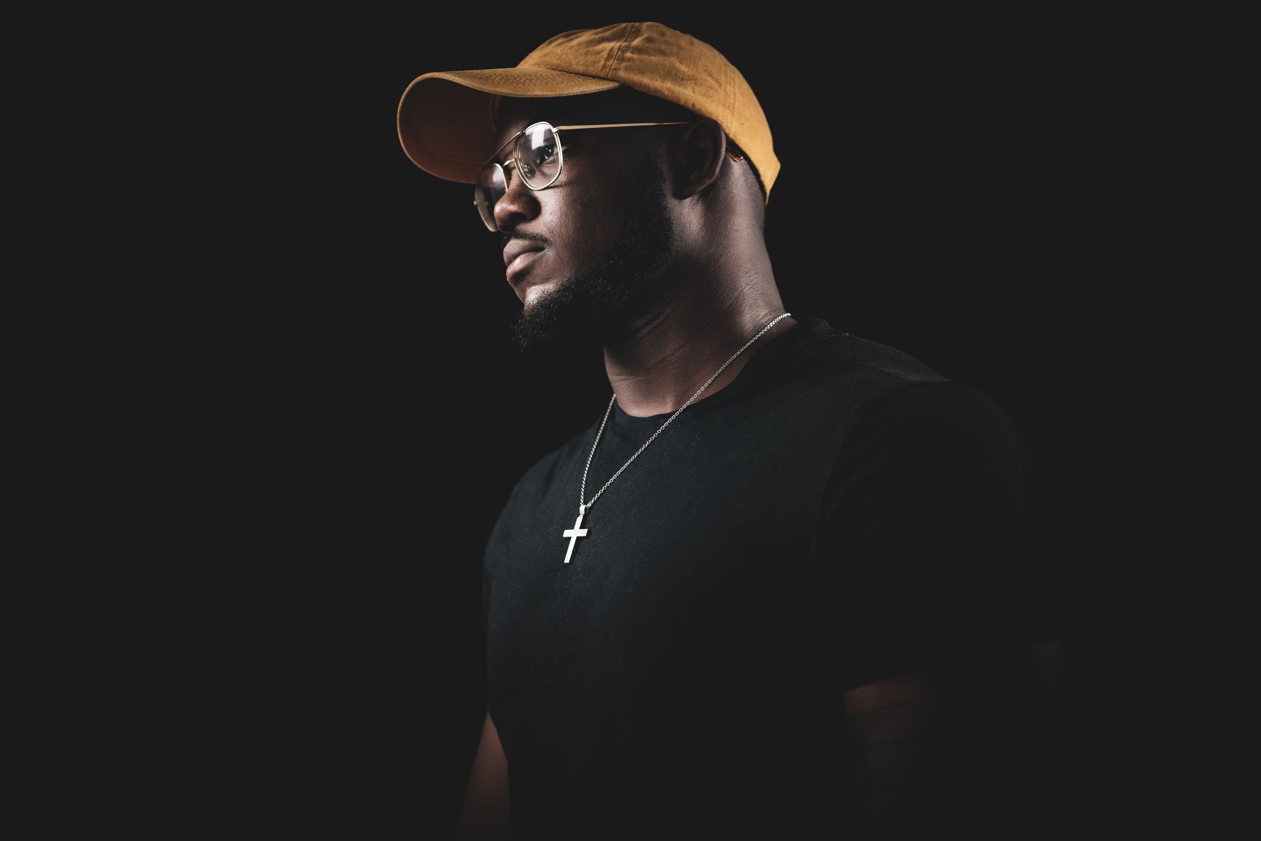 A person wearing a baseball cap and cross necklace poses against a dark backdrop.