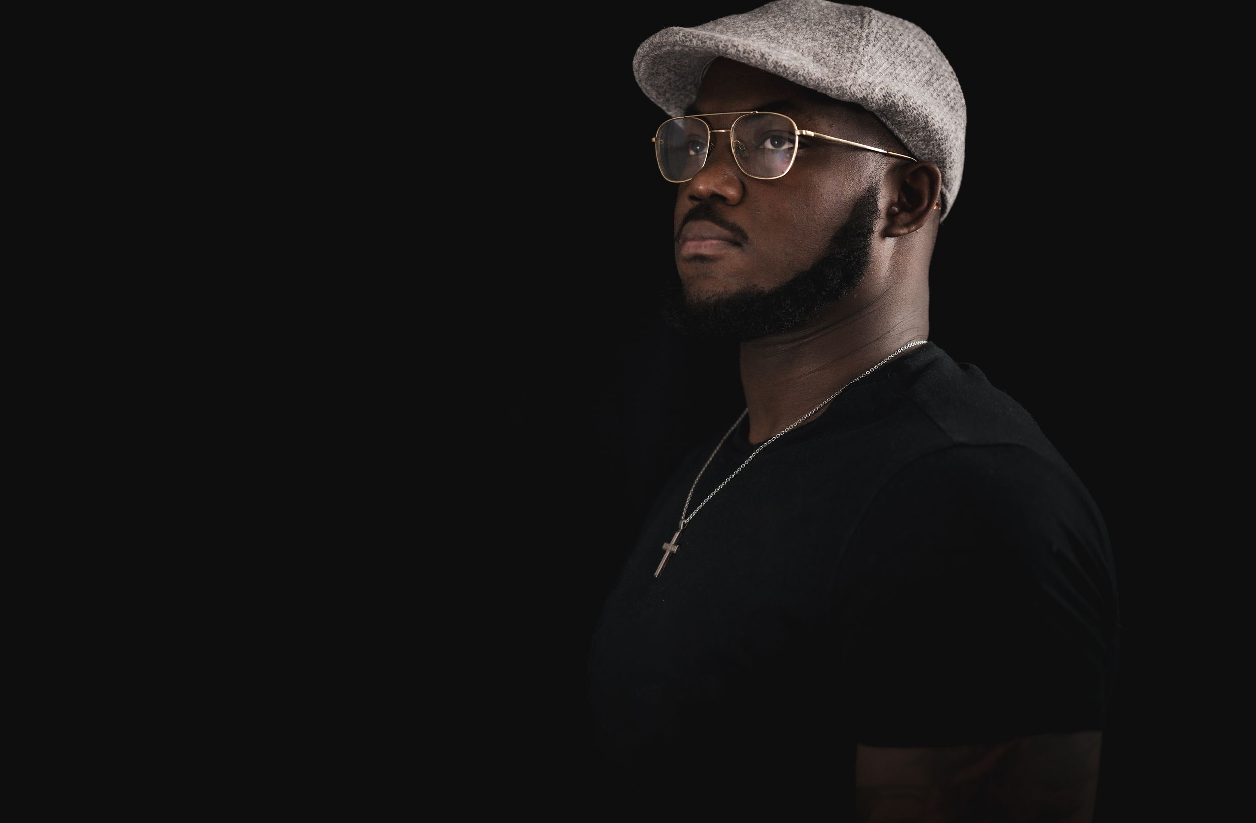 A person wearing glasses and flat cap poses against dark background in dramatic lighting.