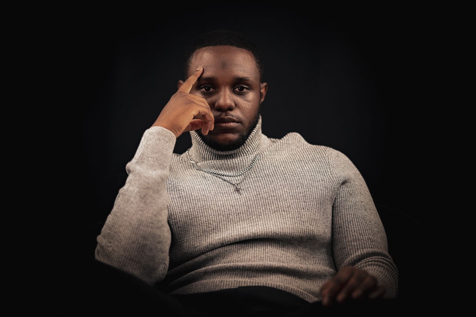 Dramatic studio portrait showing thoughtful expression in beige sweater against black backdrop.