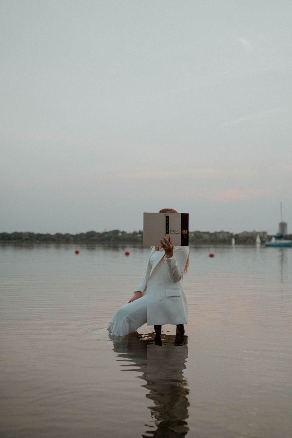 Woman in white outfit standing in lake water holding open book covering her face at dusk.