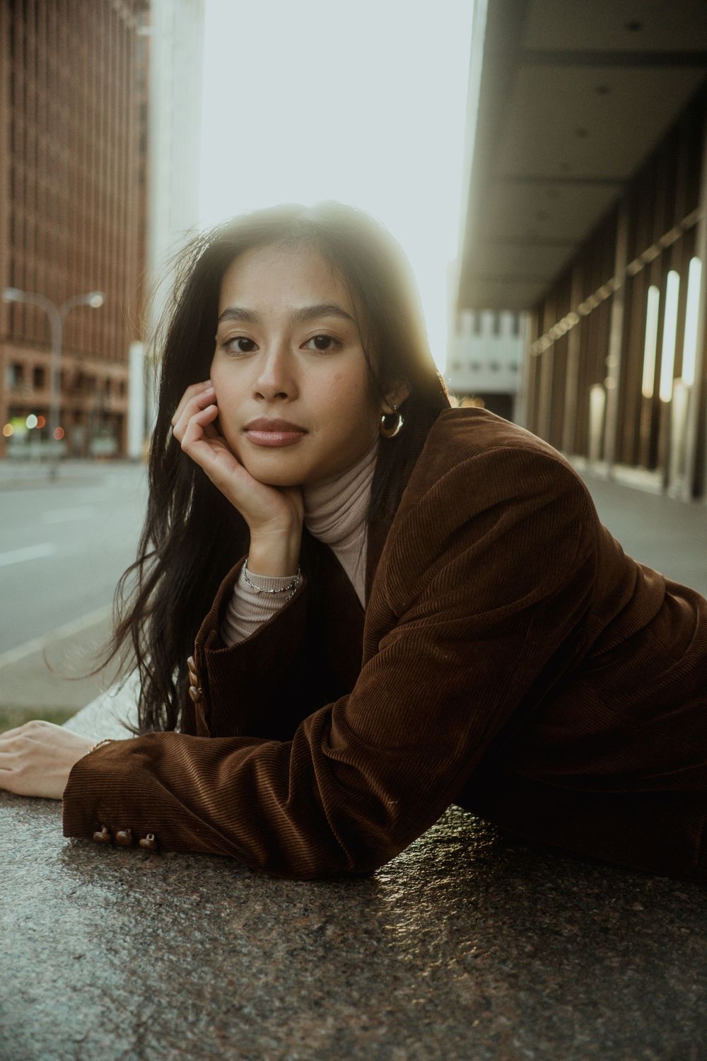 Young Asian woman in brown corduroy blazer lying on urban pavement, resting chin on hand, gazing at camera.