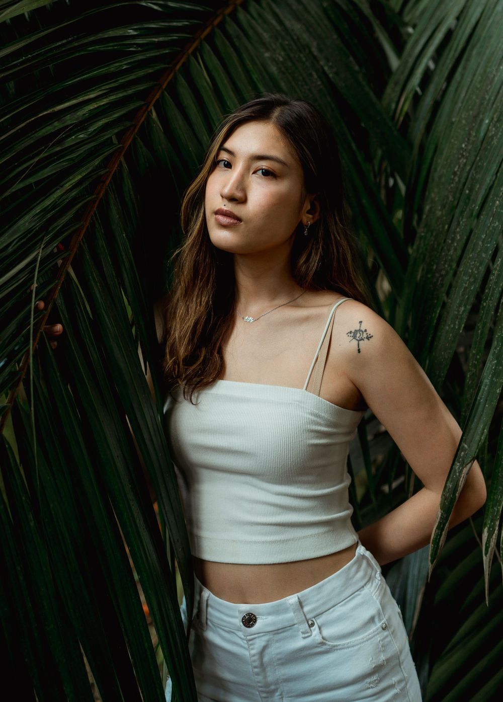 Young Asian woman in white crop top with shoulder tattoo posing among tropical palm leaves outdoors.