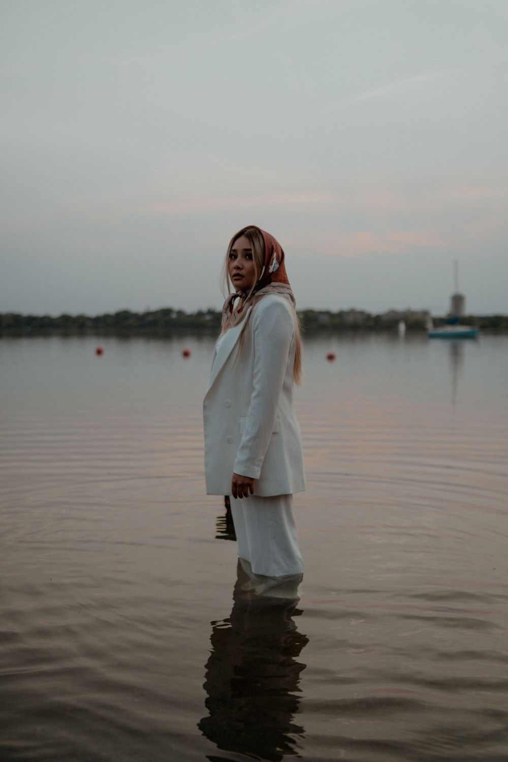 Elegant woman in white blazer and hijab standing in lake water holding sunglasses at sunset.