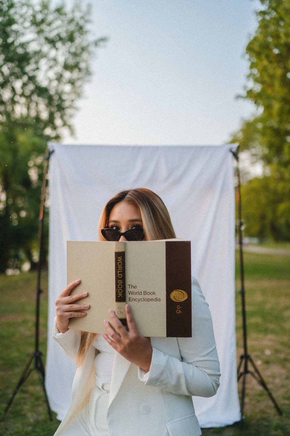 Blonde woman in white blazer holding World Book Encyclopedia outdoors near white backdrop.