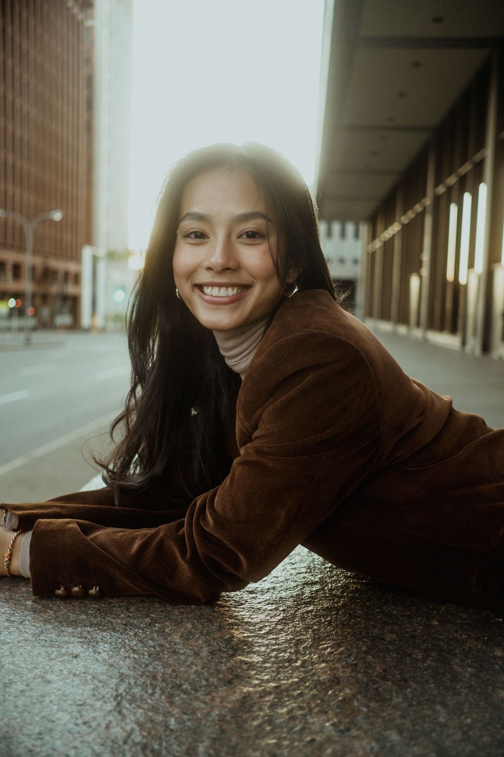 Smiling young Asian woman in brown blazer lying on city pavement with bright backlit urban background.