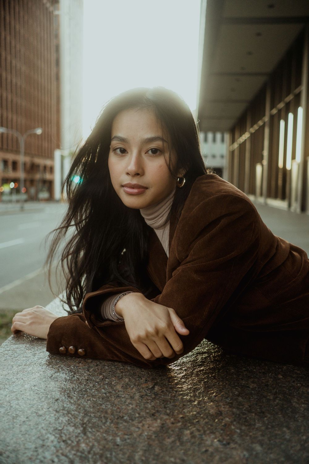 Young Asian woman in brown corduroy jacket lying on pavement, arms crossed, looking at camera outdoors.
