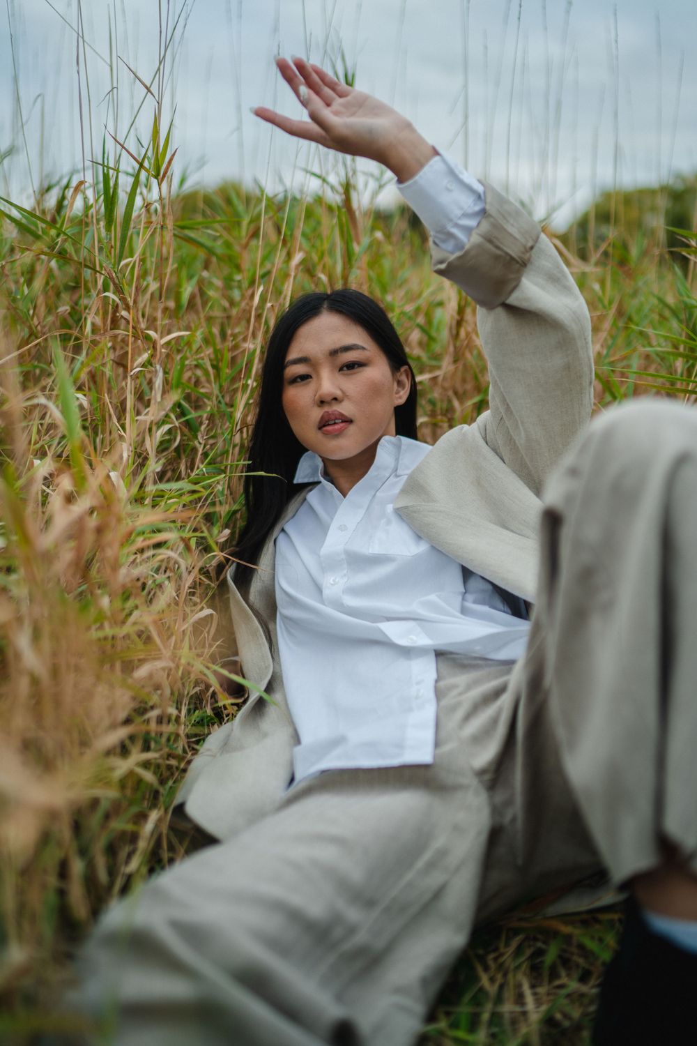 Young Asian woman in white shirt and beige suit raising hand while sitting in tall golden grass.