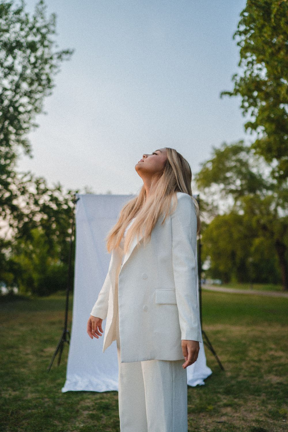 Blonde woman in white pantsuit standing outdoors with head tilted back near white backdrop.