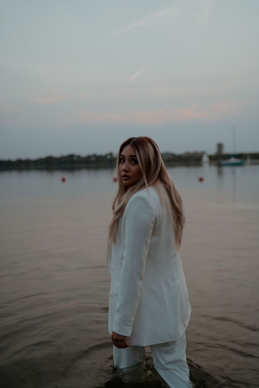 Blonde woman in white suit standing in lake water looking back over shoulder at twilight.