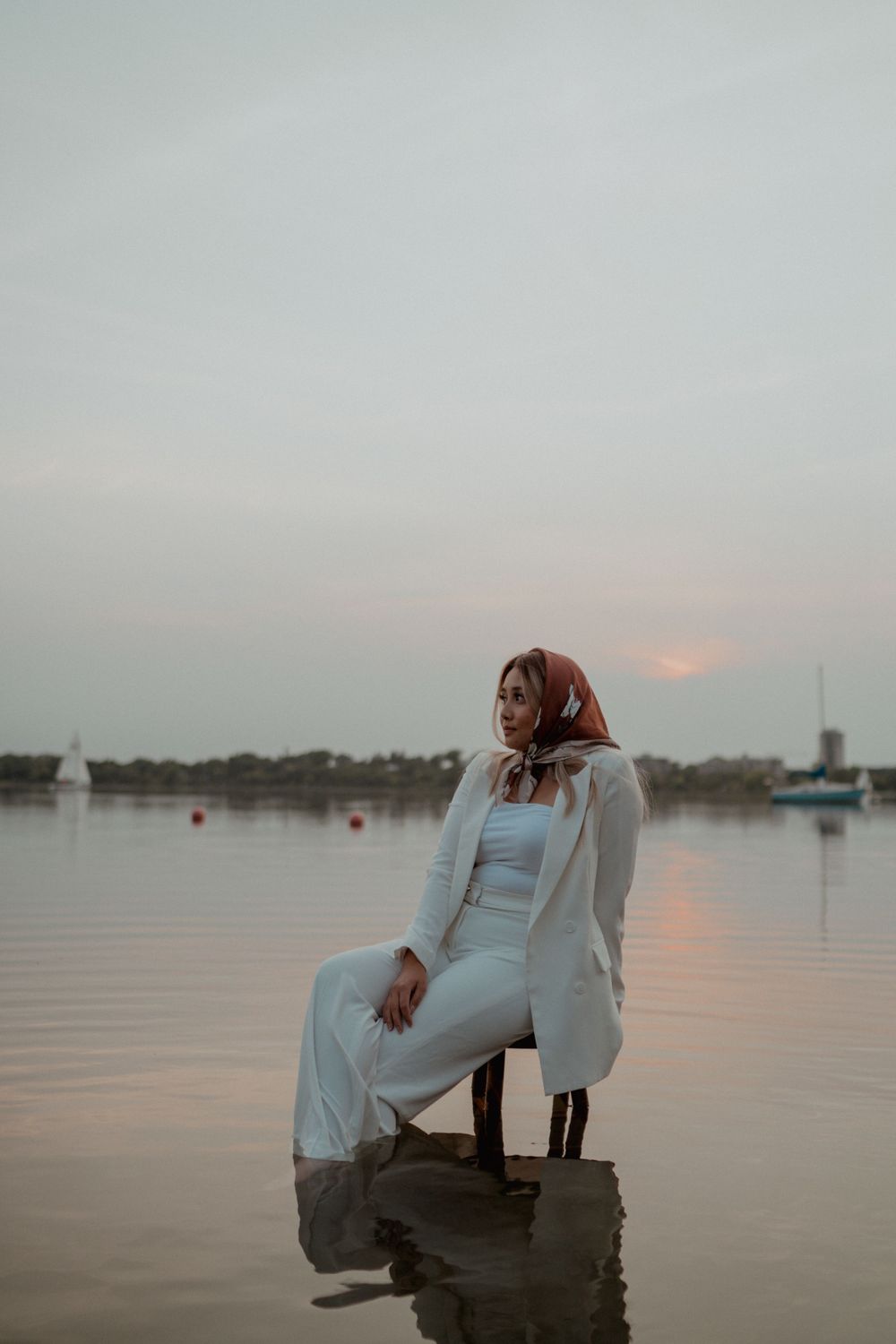 Woman in white suit and brown hijab sitting on chair in calm lake water at sunset.