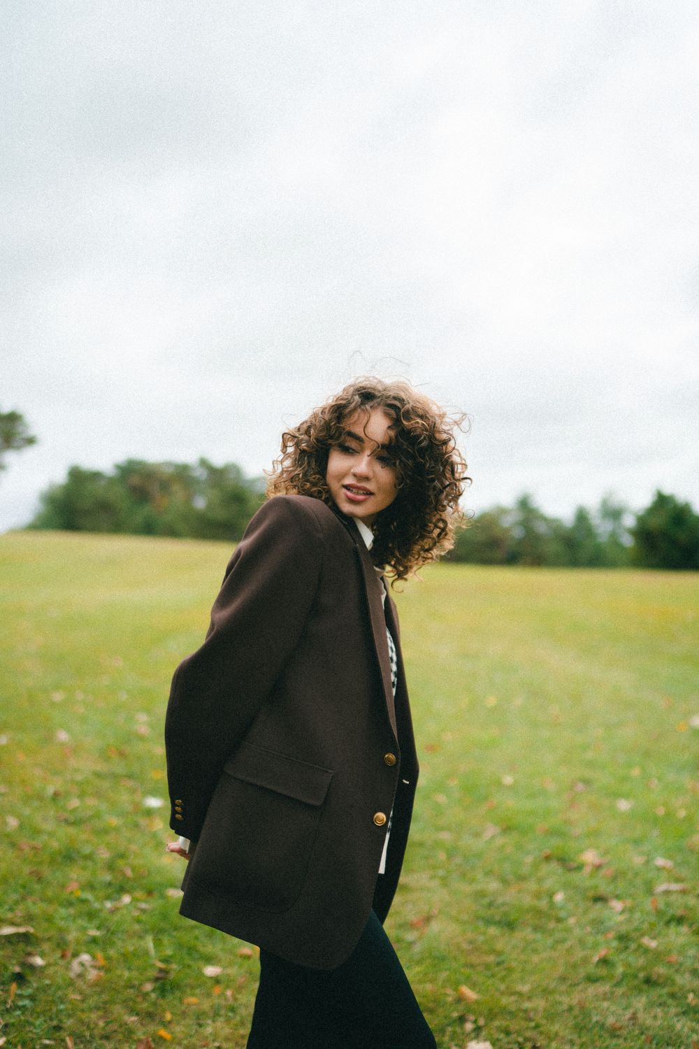 Curly-haired woman in brown double-breasted blazer smiling in open grassy field under cloudy sky.
