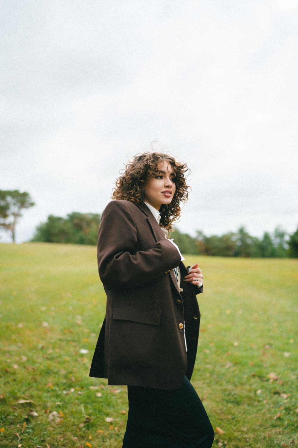Young woman with curly hair in brown oversized blazer walking in green field on cloudy day.
