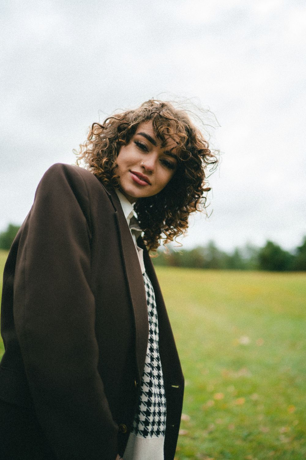 Young curly-haired woman in brown blazer and houndstooth top posing in green field outdoors.