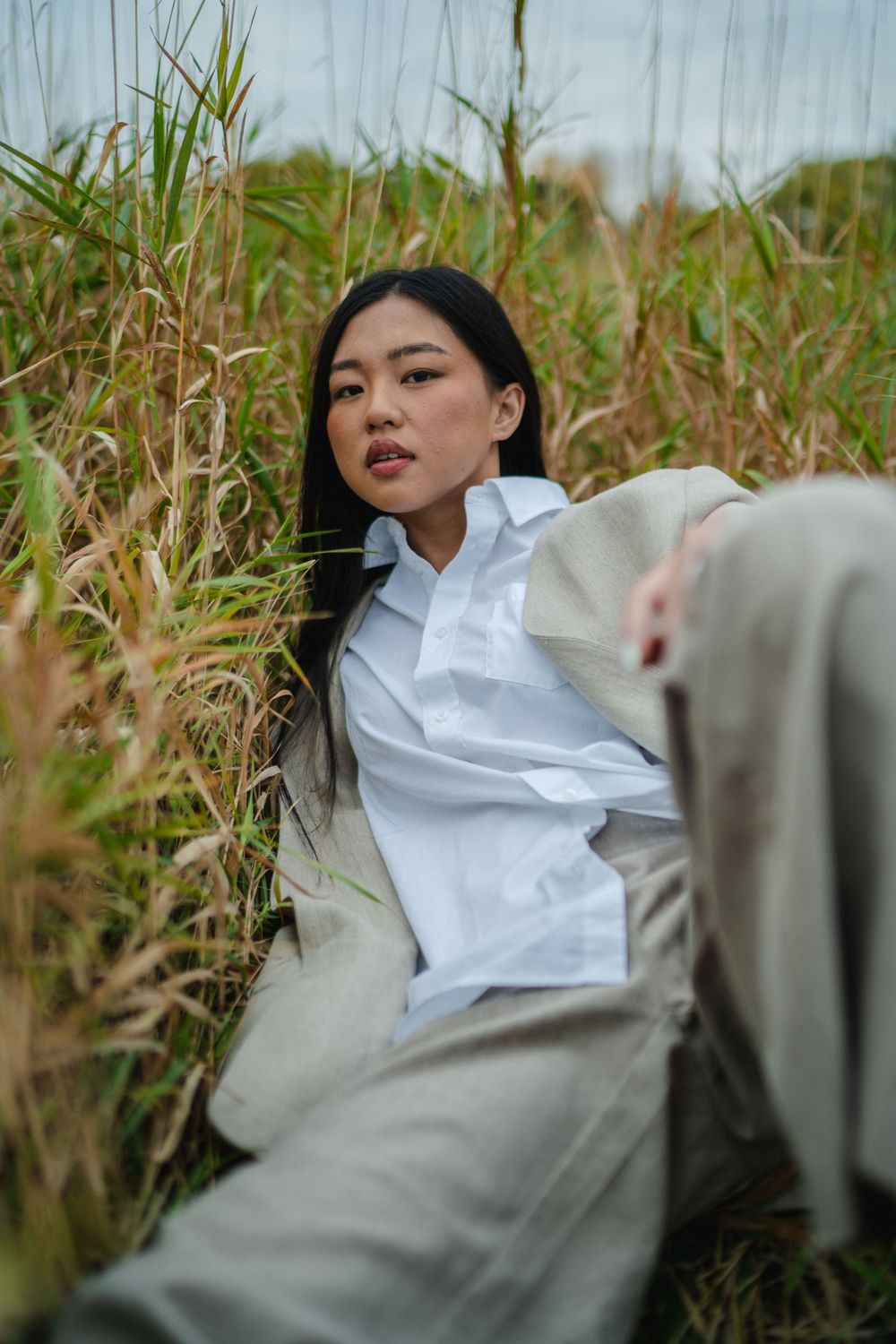 Young Asian woman in white shirt and beige suit sitting relaxed among tall golden grass in field.