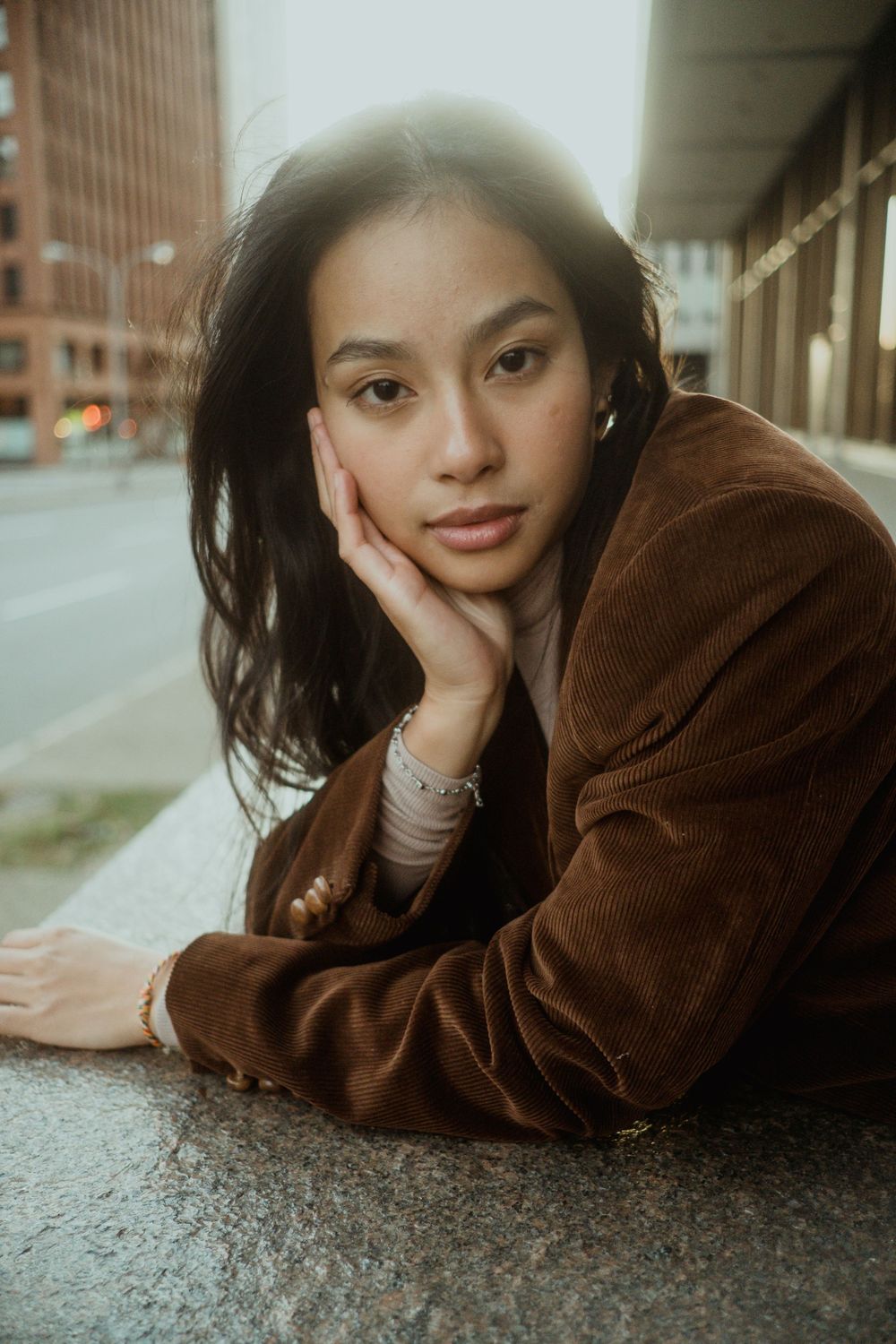 Close-up portrait of young Asian woman in brown blazer resting cheek on hand on concrete surface.