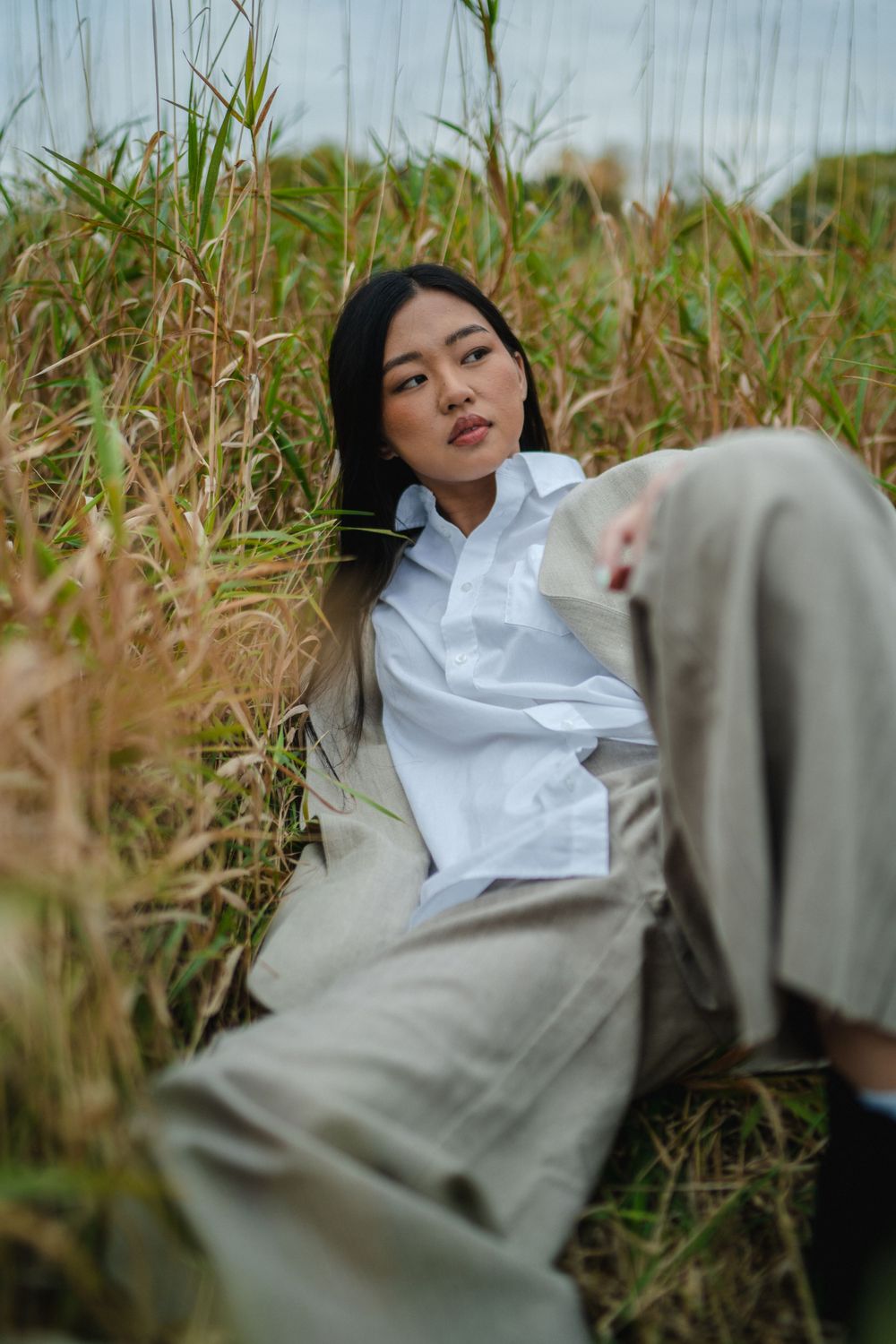 Young Asian woman in white shirt and beige trousers sitting among tall grass in an outdoor field.