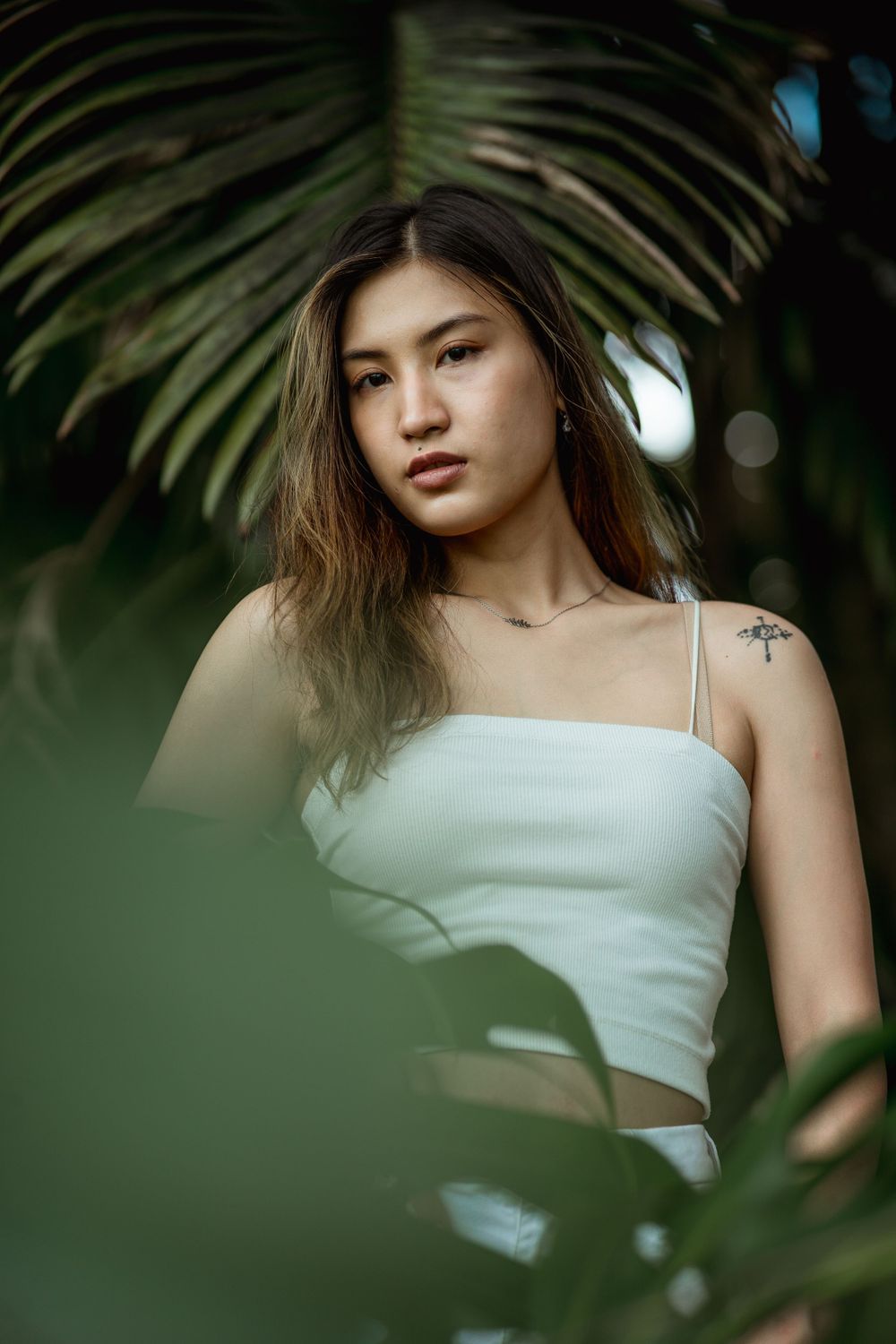 Young Asian woman in white ribbed crop top standing among lush green tropical palm leaves outdoors.