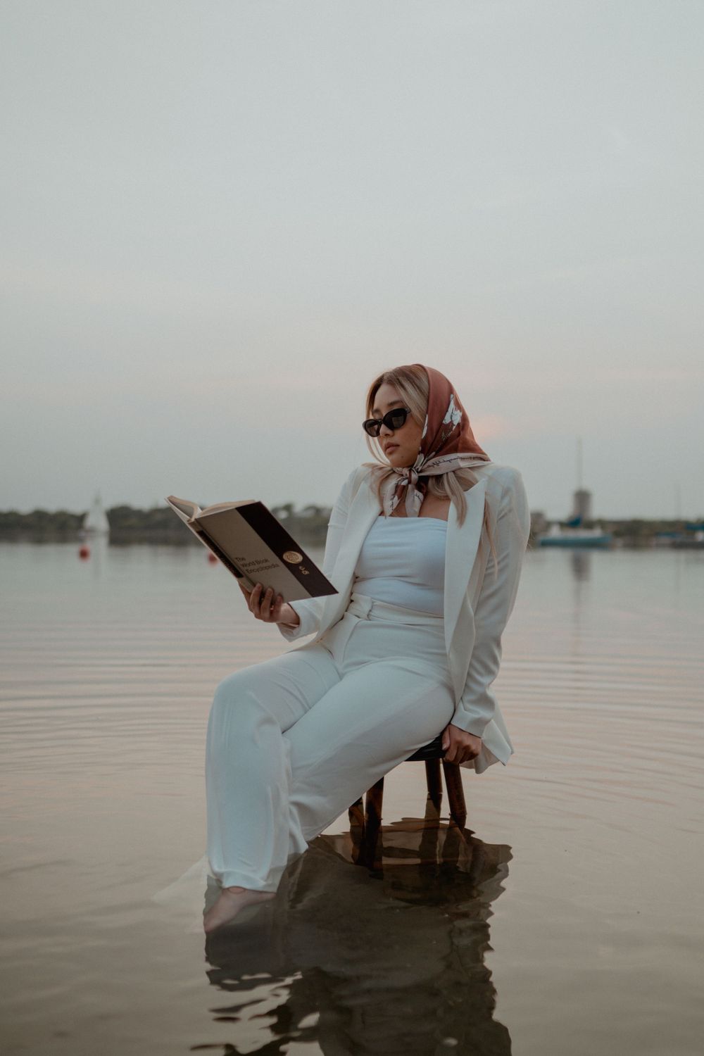 Stylish woman in white suit and hijab reading book while seated on chair in shallow lake.