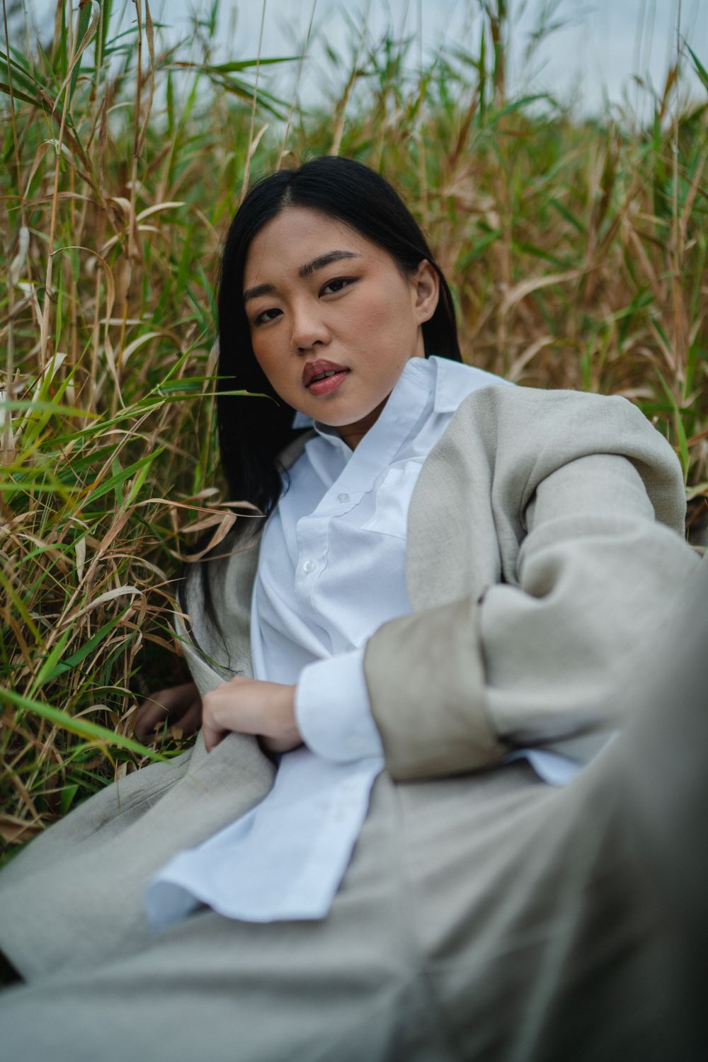 Young Asian woman in beige blazer and white shirt sitting relaxed among tall grass in open field.