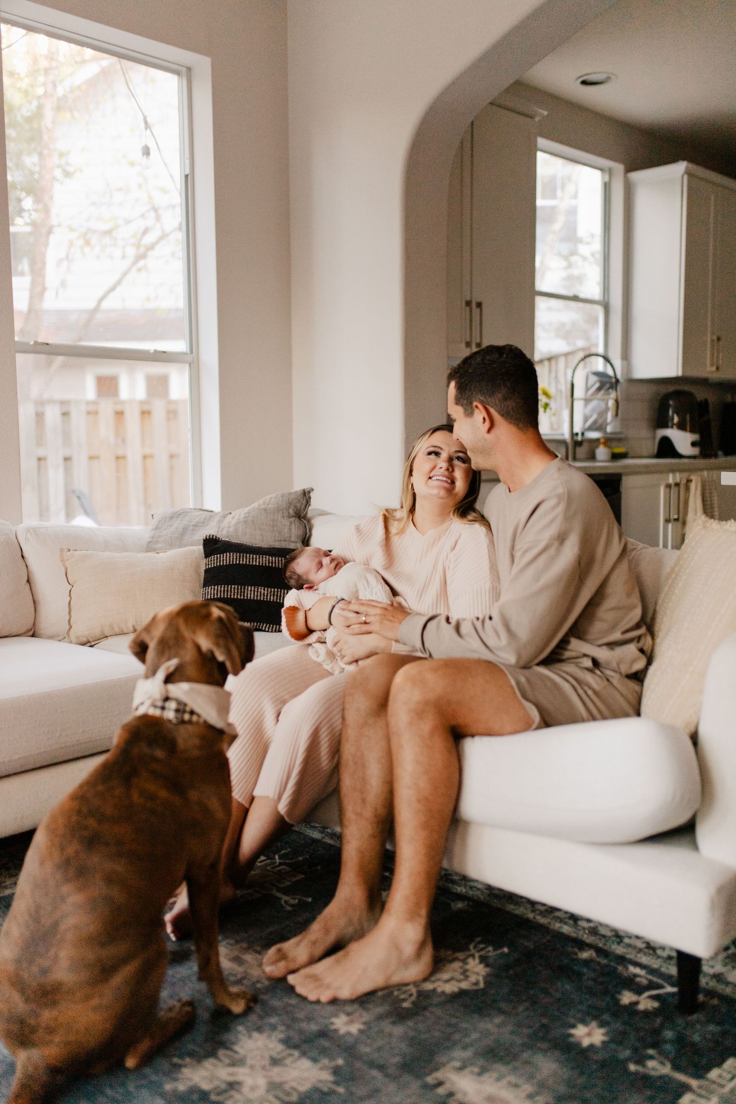 A couple cuddles with their newborn on a white couch while their dog watches attentively.