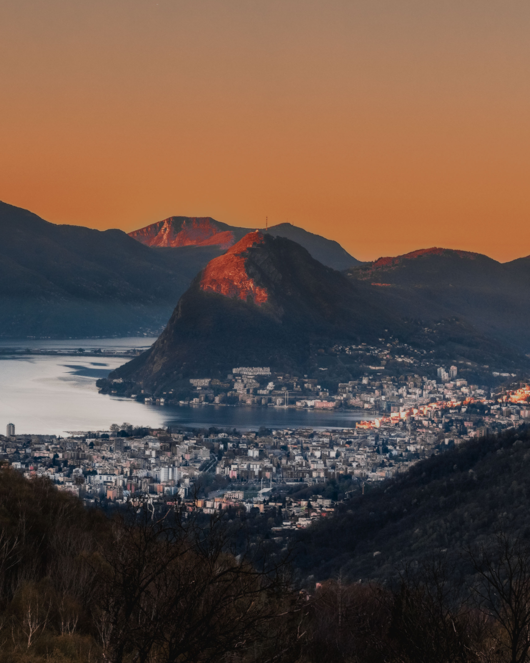 Scenic sunset view of a lakeside city nestled between mountains with a dramatic orange sky.