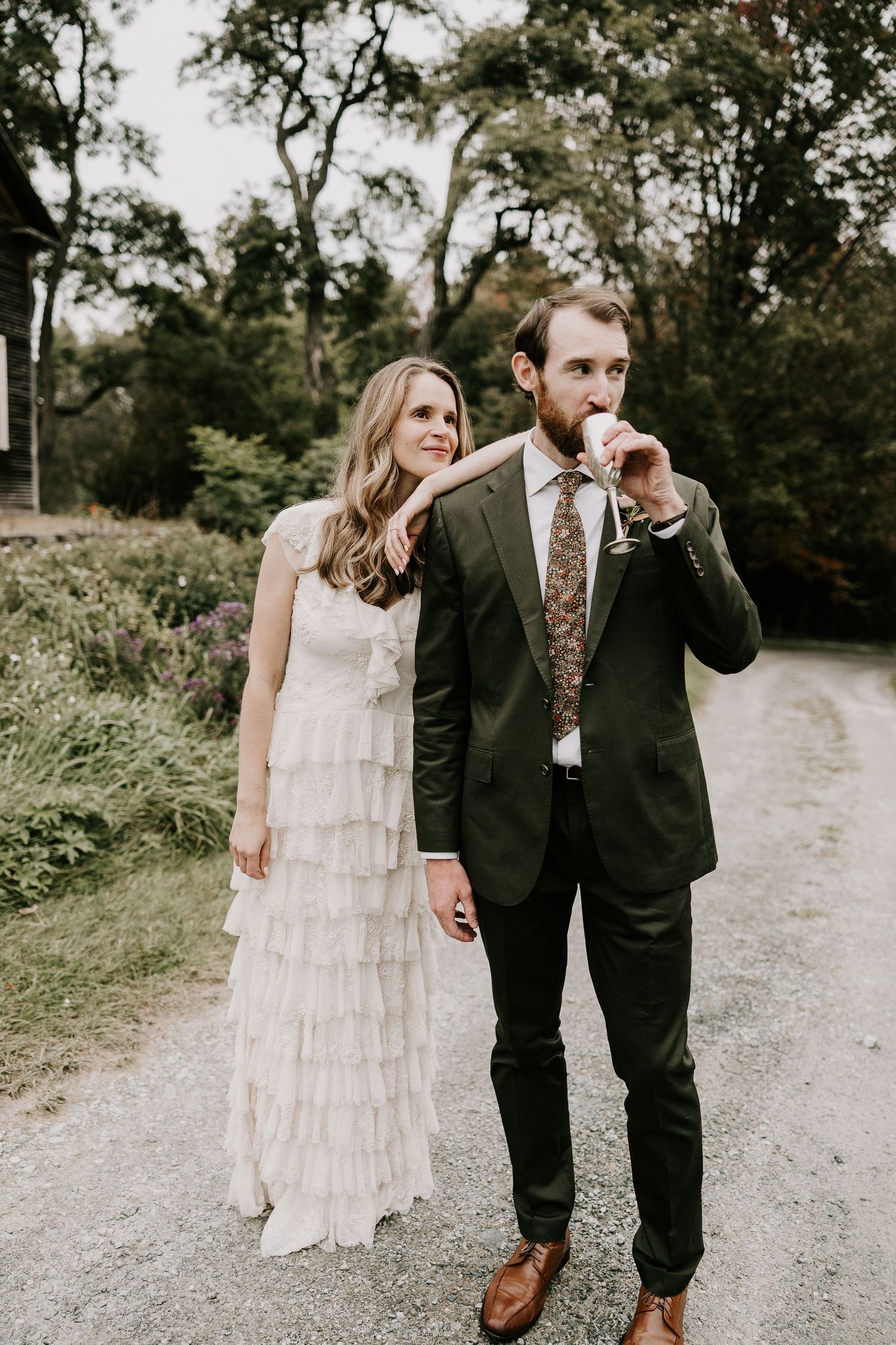 Bride in ruffled white dress stands beside groom in dark suit drinking from a cup outdoors. Newbury VT Miranda Photo & Co