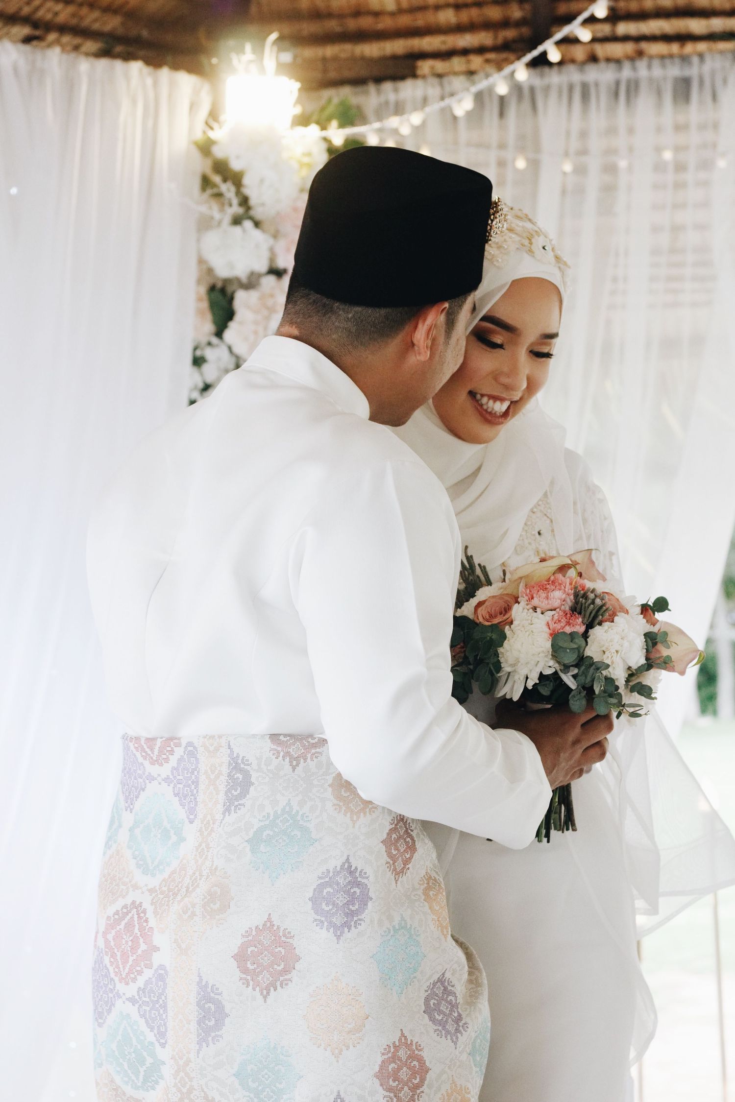 Traditional wedding couple sharing an intimate moment under thatched roof with string lights and white drapes.