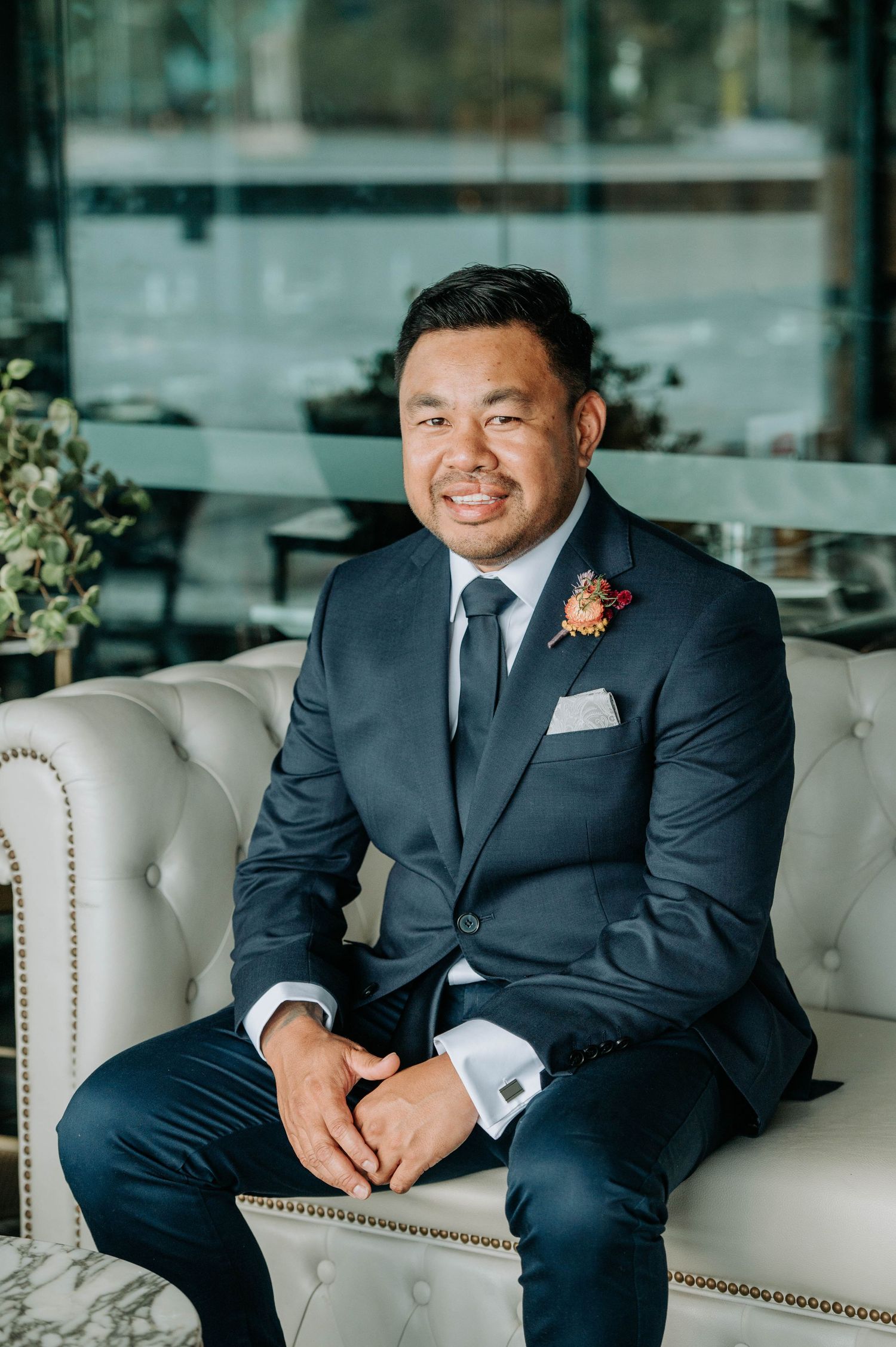 A groom sits on a luxury white leather couch in a navy suit with boutonniere.