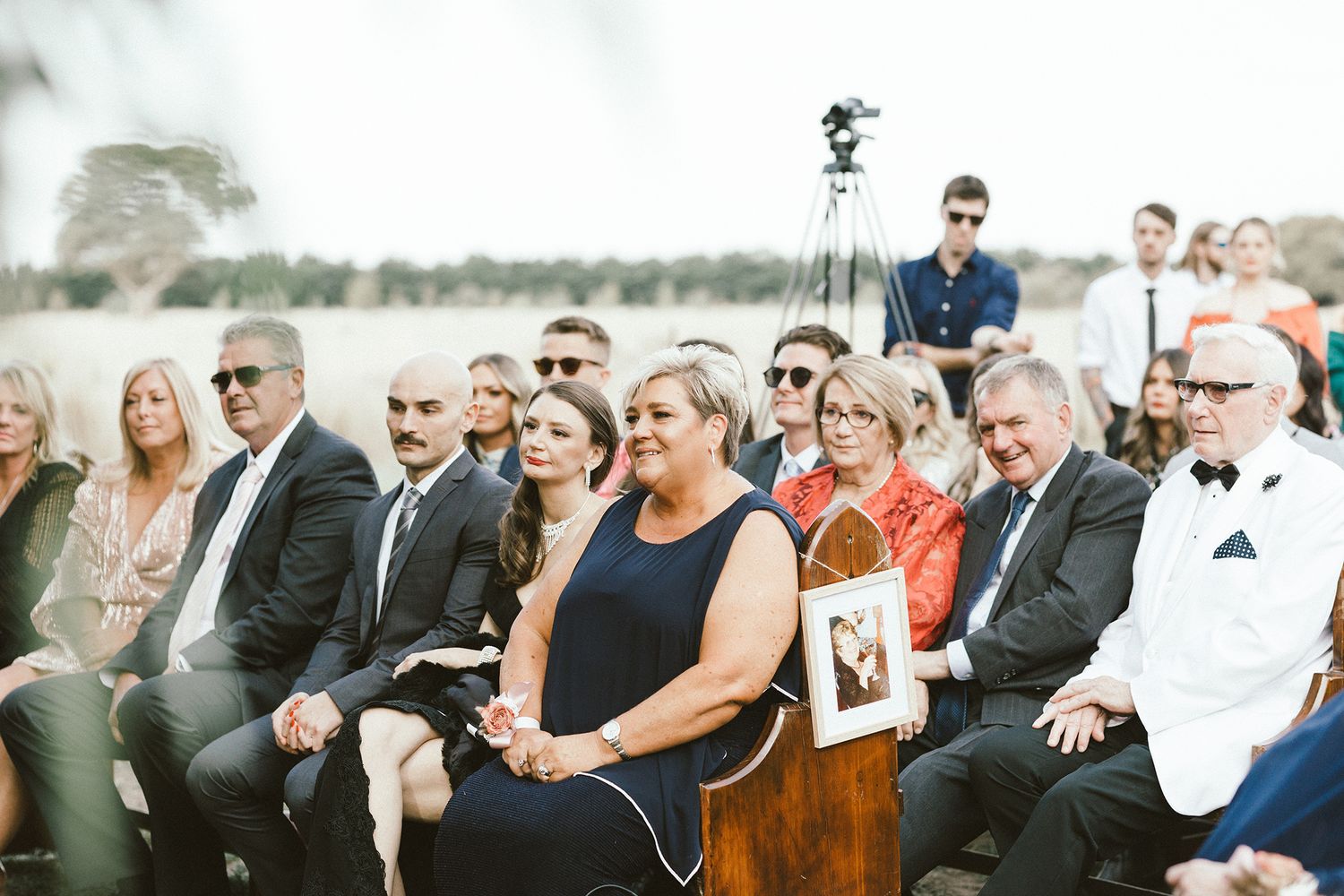 Wedding guests seated in outdoor rows watching the ceremony under trees.