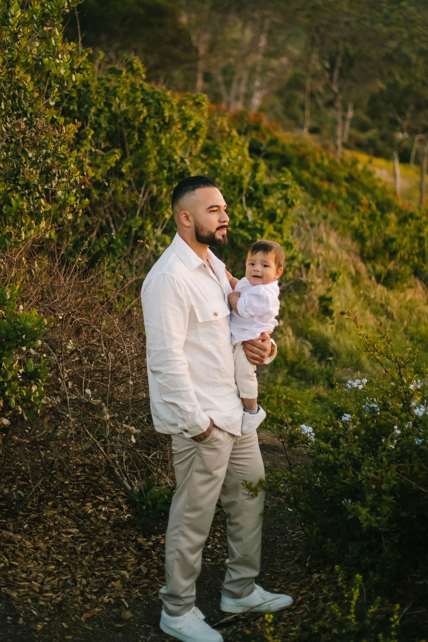 A father in white attire stands with his son against a lush green hillside background at sunset in Palos Verdes California