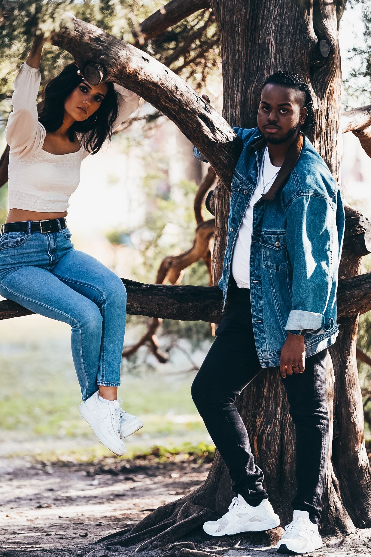 Young couple in denim posing by large tree in park, woman sitting on branch.