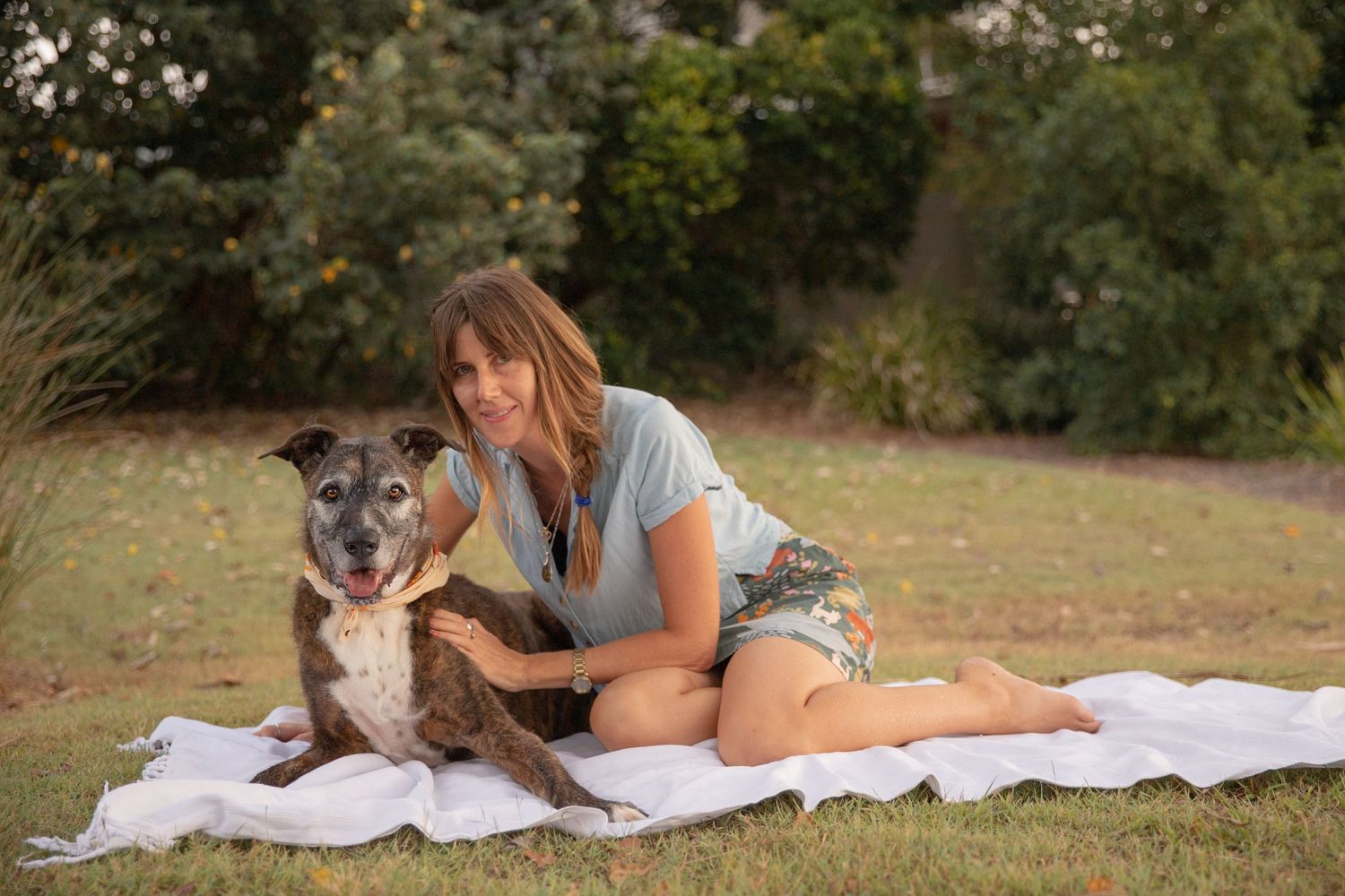 A lady and her older dog sit on a white blanket in the grass, both are looking at the camera and smiling