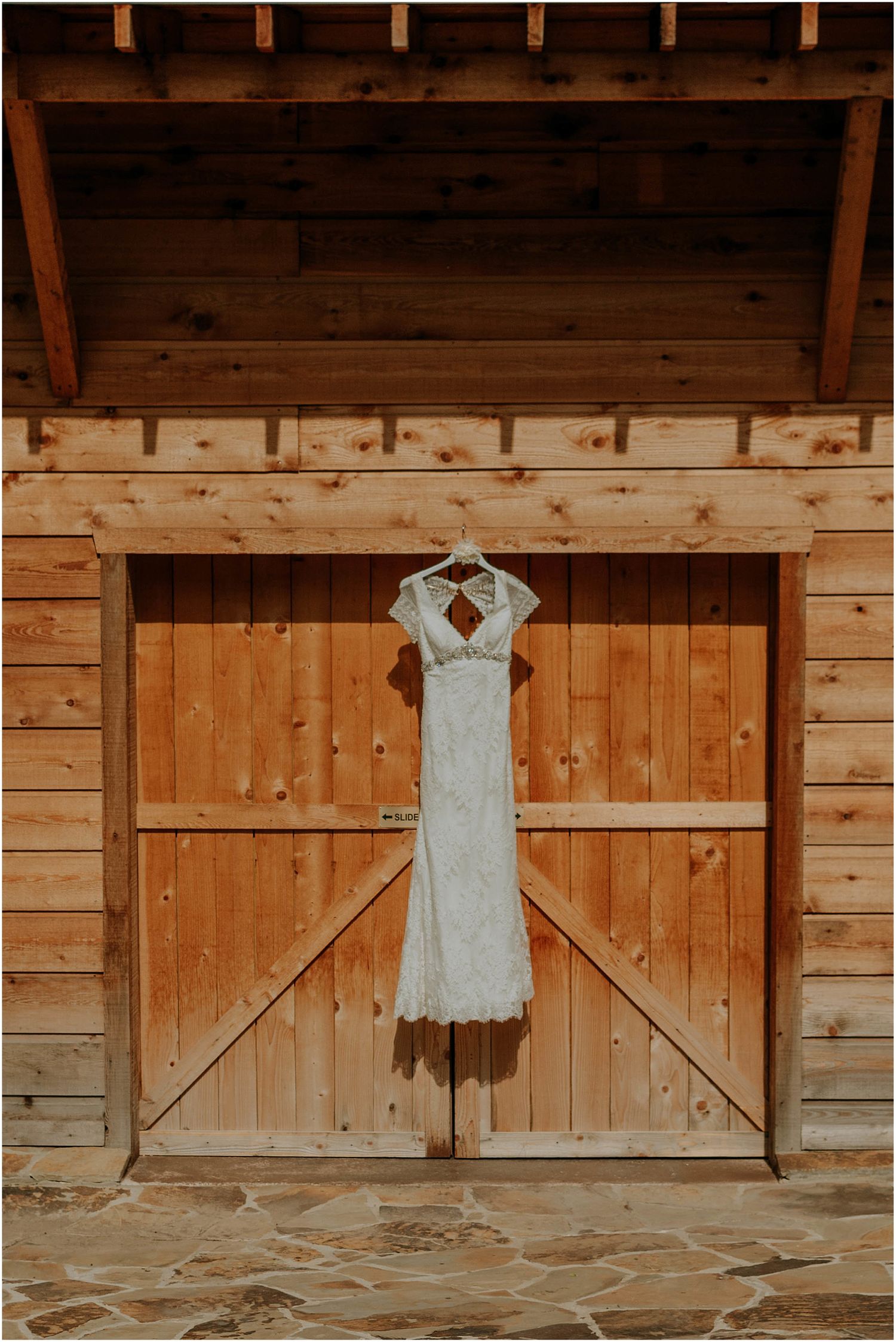 White lace wedding dress hanging on wooden barn door with stone floor.